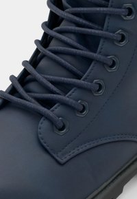 Botte en cuir bleu avec des œillets hexagonaux, un bout arrondi et une surface texturée. De longues lacets bleu foncé sont passés à travers les œillets.