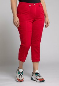 Un pantalon rouge coupé présente deux poches avant, des accents de boutons, une texture lisse et une coupe fuselée, associé à des chaussures de sport multicolores.