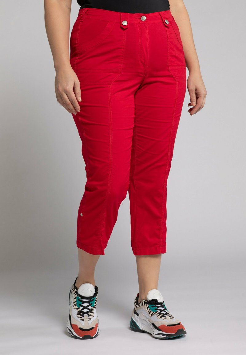 Un pantalon rouge coupé présente deux poches avant, des accents de boutons, une texture lisse et une coupe fuselée, associé à des chaussures de sport multicolores.