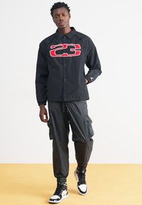 Μαύρο μπουφάν με κόκκινο λογότυπο "23", μαύρο παντελόνι cargo και μαύρα και άσπρα sneakers. Το μπουφάν διαθέτει κουμπιά, γιακά και τσέπες.