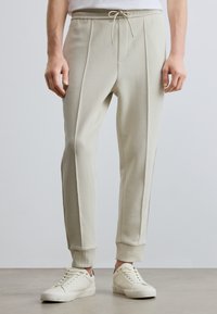 Vaaleanbeiget joggers-housut, jotka on valmistettu pehmeästä kankaasta, ja joissa on kiristysnauhavyötärö, sivutaskut sekä kapenevat nilkkapitolaiset.