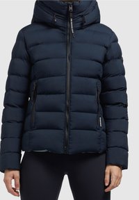 Marineblauwe pufferjas met capuchon, ritssluiting, zijzakken en doorgestikt ontwerp. Voorzien van trekkoorden en een merklabel op de mouw.