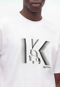 Άνδρας που φοράει ένα λευκό T-shirt με κάθετο ριγέ γραφικό με τα γράμματα "K" και μια σιλουέτα προσώπου.