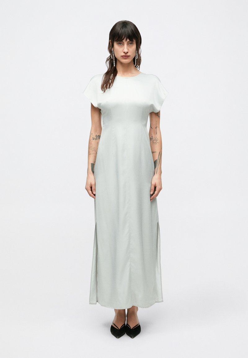 Svetlozelené saténové maxi šaty s krátkymi rozstrapkanými rukávmi, bočnými rozparkami a voľným strihom, doplnené čiernymi podpätkami so špičatou špičkou.