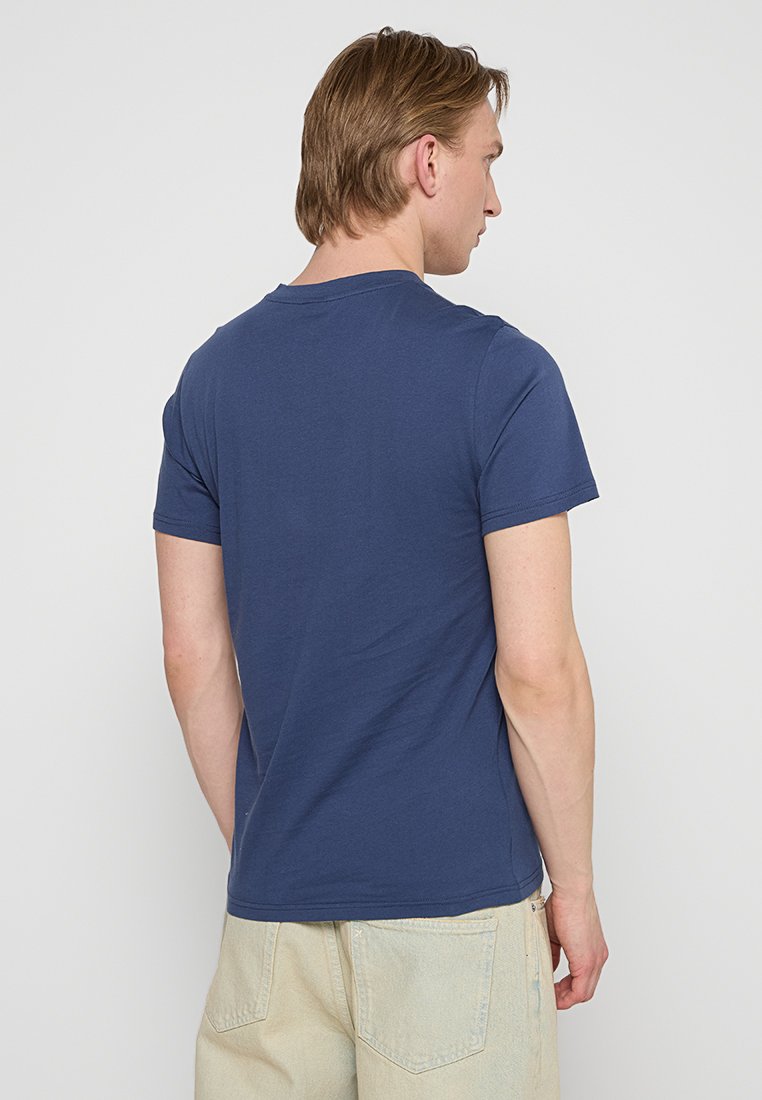 Mann mit kurzem, hellbraunem Haar, der ein schlichtes, marineblaues T-Shirt und helle beigen Hosen trägt und von hinten vor einem weißen Hintergrund steht.