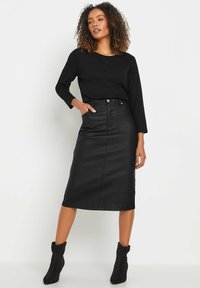 Jupe midi en cuir noir avec une couture frontale, taille haute et poches latérales, associée à un haut noir à manches longues et des bottines noires.