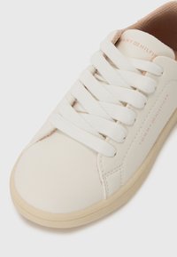 Valge nahkne sneaker, millel on sirged valged paelad, ümar nina, peene logo trükk ja beež kummipadi. Sile tekstuur ja minimalistlik disain.