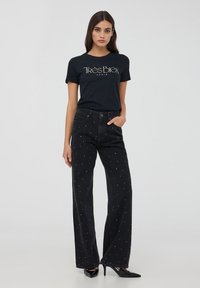 Maglietta nera con logo "Très Bien Paris", abbinata a jeans neri a vita alta con decorazioni di perline bianche e design a gamba larga.