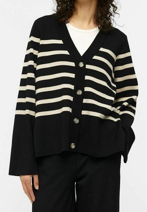 Cardigan noir avec larges rayures horizontales crème sur la moitié supérieure, boutonné devant, porté sur un top blanc et associé à un pantalon noir.