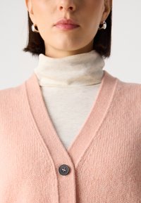 Hele roosa kardigan V-kaelusega ja tekstuurse kangaga, nööbiga sulgemine, beežist kõrge kaelusega pluusi peal. Peen ribade detail.