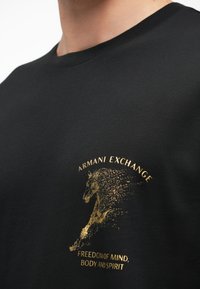 Melns kokvilnas T-krekls ar zelta drukātu grafiku ar zirgu un tekstu "Armani Exchange" un "Brīvība prātam, ķermenim un gars."