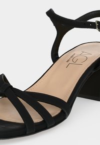 Μαύρο πέδιλο με ανοιχτά δάχτυλα με τρεις  μπροστινές λωρίδες, λουράκι στον αστράγαλο με αγκράφα, μπεζ επενδυμένο πάτο και χαμηλό τετράγωνο τακούνι.