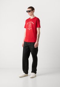 Rød bomulds t-shirt med "GANT SPORTSWEAR" print, tilsat sorte denimjeans og hvide sneakers. Tilbehøret inkluderer sorte solbriller.