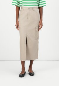 Jupe cargo beige avec une fente sur le devant, plusieurs poches et une ceinture à fermeture avec bouton. Le modèle est associé à des ballerines noires et un haut rayé vert.