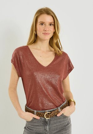 Mujer con cabello rubio largo que lleva una blusa brillante de color óxido con escote en V, jeans grises, cinturón con estampado de leopardo, pendientes de aro dorados y una pulsera dorada.