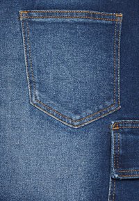 Detailní záběr modré džínoviny s našitým zadním kapsou a částí menší kapsy s oranžovým prošíváním.