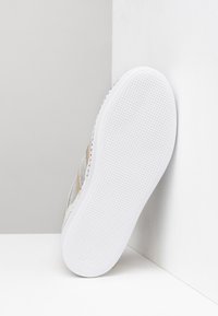 Sneaker con una toma in tessuto argentato e dettagli beige, suola in gomma bianca texturizzata e un design slanciato senza lacci visibili.