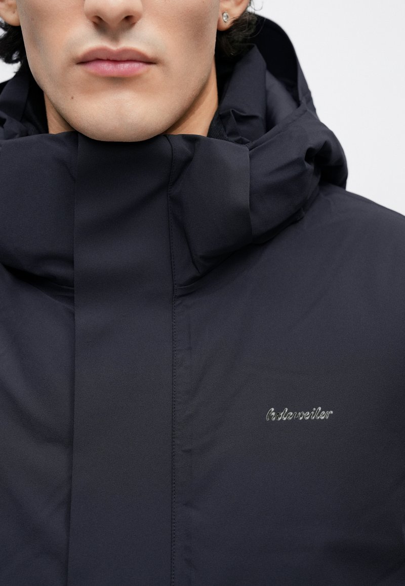 Navy blue jacket with a high collar, featuring a silver logo embroidered on the chest. The material has a smooth texture and sleek design.