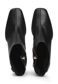 Botines de cuero negro hasta el tobillo con punta cuadrada, que presentan detalles de cremallera dorada y costuras visibles. Textura suave y silueta esbelta.