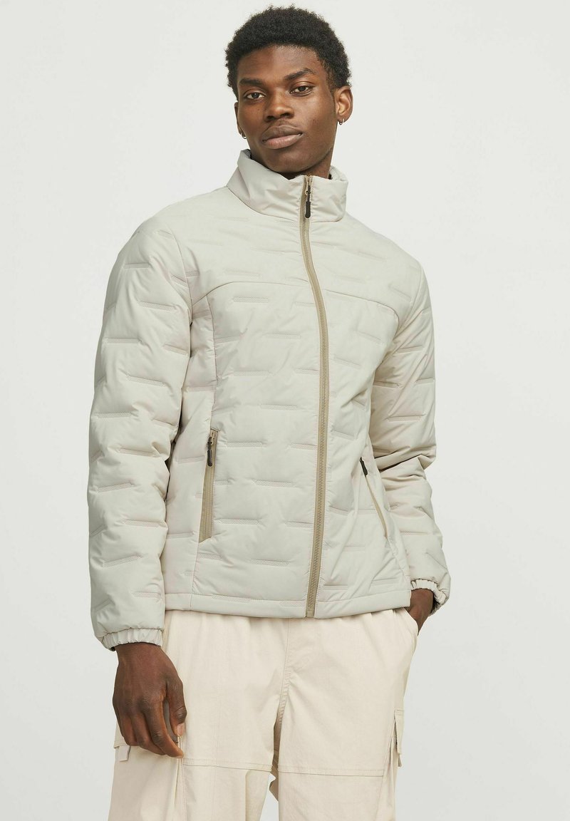 Veste isolante gris clair avec col haut, fermeture éclair intégrale, coutures diagonales et deux poches latérales. Fabriquée en tissu lisse et doux.