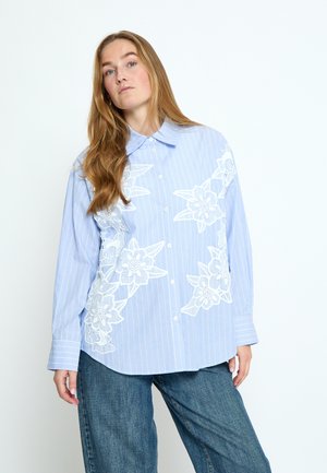 Vrouw draagt een blauw gestreept overhemd met witte bloemrijke borduursels, gecombineerd met loszittende blauwe spijkerbroek, staand tegen een witte achtergrond.