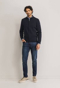 Marineblauer Zip-Up-Pullover aus Strickstoff, mit hohem Kragen und gerippten Bündchen, kombiniert mit dunklen Jeans und beigen Sneakers.