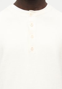 Κοντινό πλάνο ενός ατόμου που φοράει μια εκρού υφασμάτινη μπλούζα henley με τέσσερα κρεμ κουμπιά στον πάτο.