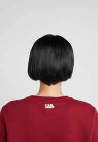 Burgundyröd sweatshirt med rund halsringning, med en vit "KARL LAGERFELD" logga på ryggen. Rakt svart bobhår synligt bakifrån.