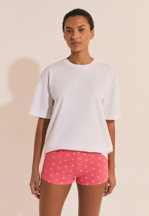 Frau steht vor einfachem Hintergrund und trägt ein lockeres weißes T-Shirt sowie rosa Shorts mit kleinen weißen Blumenmustern.