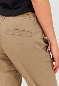 Pantalons khaki à texture lisse, comportant une poche arrière et une poche latérale. Une étiquette de marque est visible sur la ceinture.