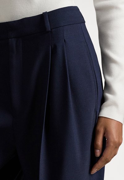 Κοντινό πλάνο ατόμου που φοράει μπλε σκούρο πλισέ παντελόνι και λευκό μακρυμάνικο τοπ, με το χέρι ακουμπισμένο κοντά στο ισχίο μπροστά σε απλό φόντο.