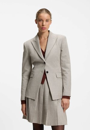 Kvinde iført en beige ternet blazer og matchende plisseret nederdel over en bordeaux top, stående mod en ensfarvet hvid baggrund.