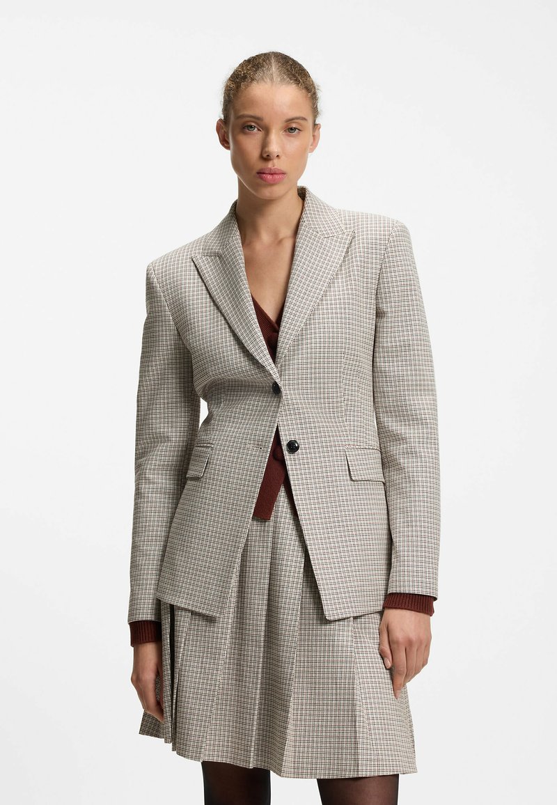 Femme portant un blazer à carreaux beige et une jupe plissée assortie sur un haut bordeaux, debout devant un fond blanc uni.