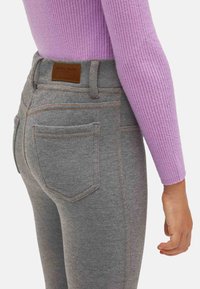 Jeans grises de cintura alta con costuras naranjas, que presentan dos bolsillos traseros y un parche de cuero marrón de la marca. Combinados con un top de canalé morado.