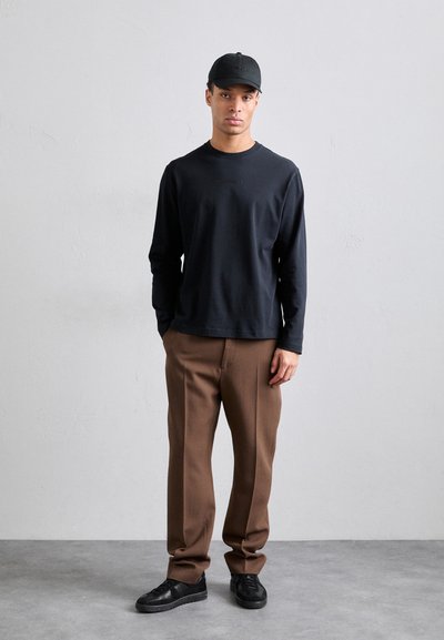 Chemise noire à manches longues, pantalon brun sur mesure et baskets noires. La casquette est noire avec un logo discret. Design simple, coupe décontractée.