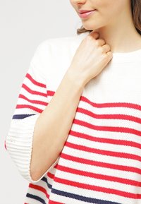 Suéter blanco con rayas horizontales rojas y marinas, cuello redondo y corte holgado. Textura de punto con puños de manga enrollados.