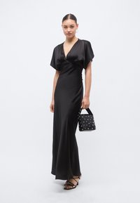 Μακρύ μαύρο φόρεμα από σατέν με βαθύ V-λαιμόκοψη, κοντά φαρδιά μανίκια και εφαρμοστή μέση. Συμπληρώνεται από μια μικρή μαύρη τσάντα με τρουκς.