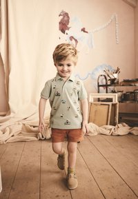 Polo vert clair avec broderie multicolore sur le thème de la ferme, associé à un short marron. L'enfant porte des chaussettes vertes et des chaussures marron sans lacets.