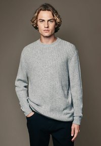 Light gray ribbed sweater with a crew neckline and long sleeves, made of soft fabric. Paired with dark trousers, against a neutral background.