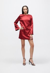 Piros szatén miniruha hosszú harang ujjakkal, kerek nyakkivágással és sima textúrával. Fekete pántos magas sarkú cipővel viselve.