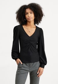 Black long-sleeve top with a V-neck and gathered detail at the front. Features puffed sleeves and smooth texture. Paired with houndstooth patterned pants.