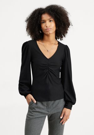 Black long-sleeve top with a V-neck and gathered detail at the front. Features puffed sleeves and smooth texture. Paired with houndstooth patterned pants.