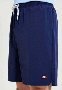 Maritiemblauwe sportshorts met elastische tailleband, witte trekkoord met het merk "ellesse" en een klein kleurrijk logo dicht bij de zoom.