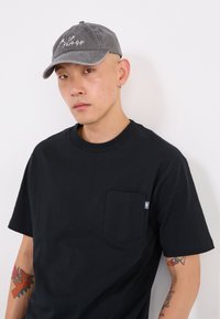 T-shirt en coton noir avec une poche sur la poitrine, associé à une casquette grise brodée avec le texte "LA PLAGE". Peau visible, ornée de tatouages.