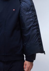 Mornarska zip-up jakna z mehkobo in teksturo tkanine ter gladkim, sijočim podlogom. Žameti na koncih rokavov in na robu, z majhnim logotipom zastave na prsih.