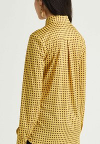 Camicia gialla con collo a revers, caratterizzata da un motivo di piccoli cerchi marroni. Maniche lunghe e dettaglio plissettato sul retro. Tessuto dalla texture liscia.