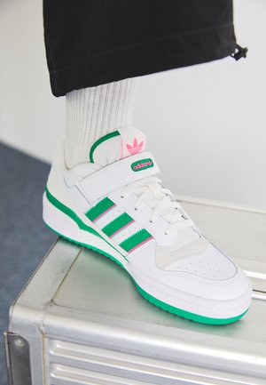 Zapatilla blanca con acentos en verde y rosa, tres rayas verdes y una puntera texturizada. Cuenta con una correa de Velcro y suela de goma.