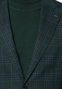 Zielona marynarka w kratę o gładkiej teksturze, z ciemnozielonym golfem pod spodem. Do zauważalnych detali należą oznakowana klapa i guzik.