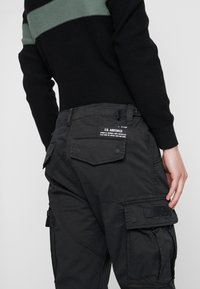 Sorte cargo bukser med en struktureret finish, der har flere lommer og et "ALPHA" logo-patch, parret med en sort og grøn stribet sweater.