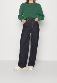 Grön ulltröja med puffärmar, i kombination med hög midja mörka denimjeans och krämfärgade skor med tjock sula, kompletterat med ett svart bälte.